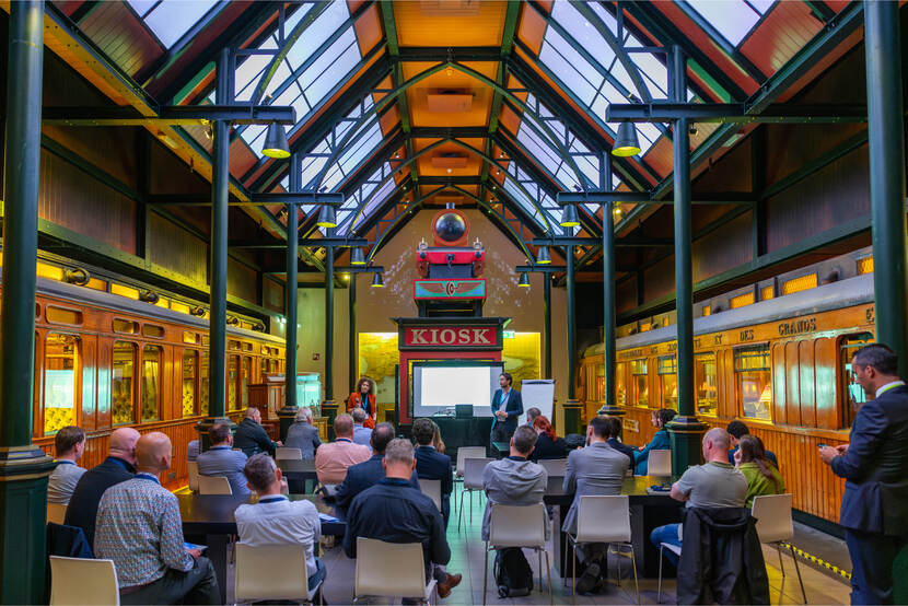 Presentatie aan een groep mensen in een historische hal in het Spoorwegmuseum met een glazen dak en treinwagons aan weerszijden, met een bord ‘KIOSK’ op de achtergrond.