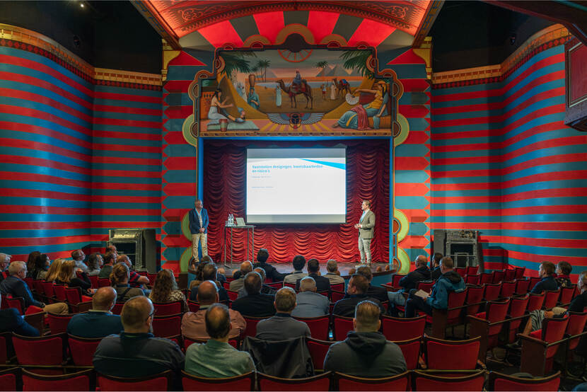 Presentatie in een kleurrijk theater in het Spoorwegmuseum met rood-blauwe gestreepte muren en een rijk gedecoreerd podium, voor een publiek in rode stoelen