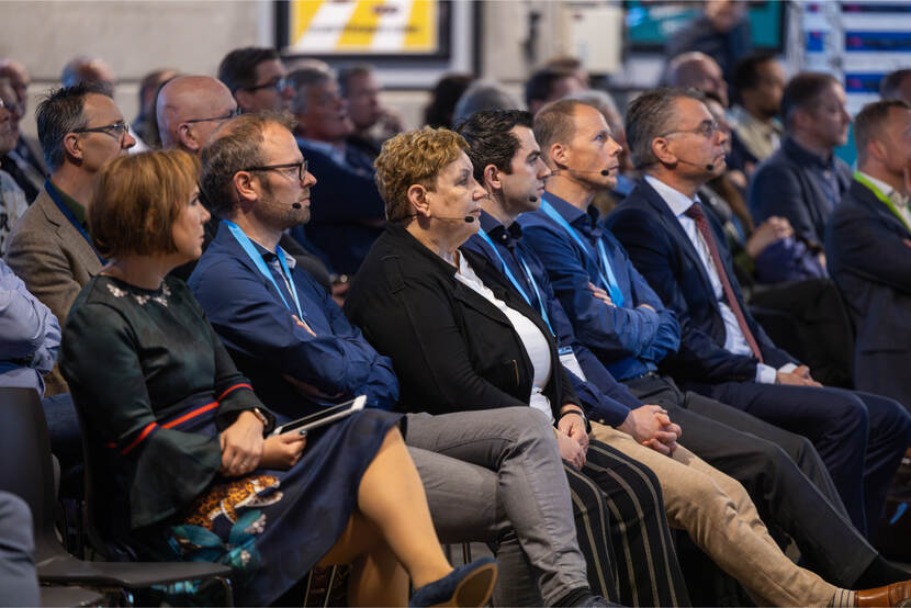 Groep aandachtige congresdeelnemers, waaronder moderator Jessica Maas, met microfoons, zittend in rijen tijdens een presentatie in de grote zaal.