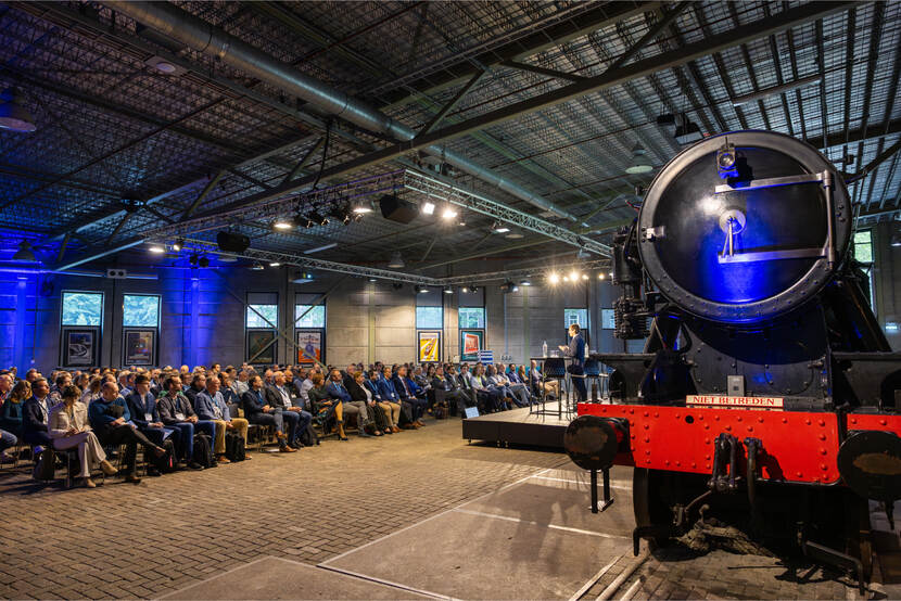 Spreker van het Europees parlement presenteert over het Europees cybersecuritybeleid en specifiek de NIS2 aan een groot publiek in de grote zaal van het Spoorwegmuseum met een stoomlocomotief op de voorgrond.