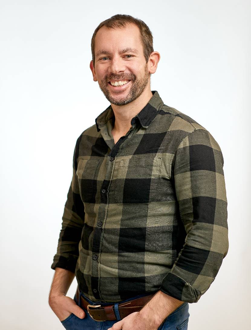 Kelvin heeft kort bruin haar en een lichte baard. Hij glimlacht vriendelijk naar de camera. Hij draagt een groen-zwart geruite blouse en heeft zijn handen ontspannen in zijn zakken. De achtergrond is licht en neutraal.