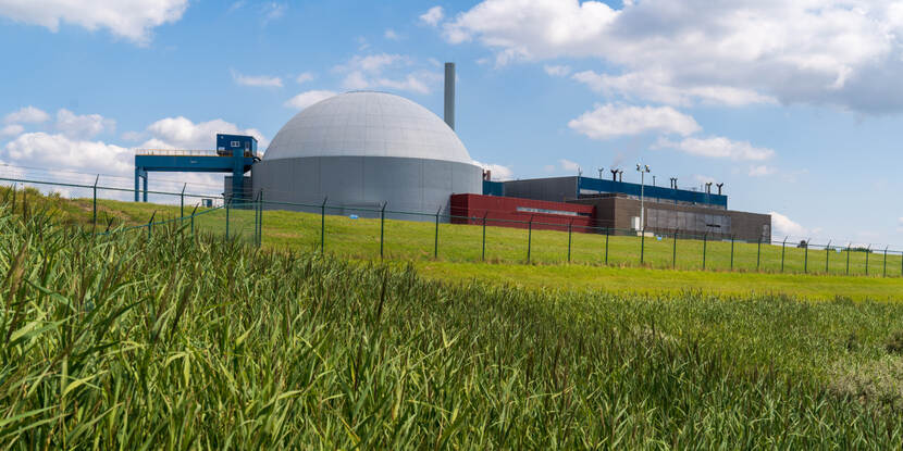 Kerncentrale met een koepelvormig reactorgebouw en industriële bijgebouwen, omgeven door een hekwerk en grasland onder een deels bewolkte hemel.
